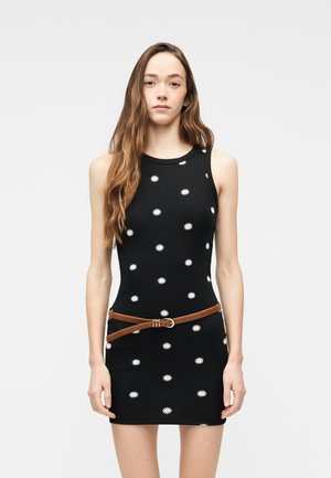 Ung kvinde iført en ærmeløs sort kjole med hvide margueritmønstre og et brunt bælte, stående mod en ensartet lys baggrund.