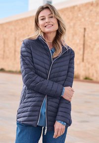 Marineblauwe gewatteerde pufferjack met een hoge kraag en ritssluiting, met een gladde textuur en een schuin stikselpatroon, gedragen met een denimhemd.