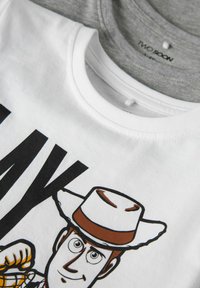 Valge lühikeste varrukatega t-särk, millel on koomiksitegelane, kes kannab cowboy mütsi ja millel on suur trükitud must "PLAY" tekst. All on nähtav hall t-särk.