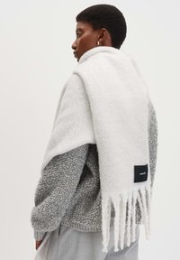 Sciarpa bianca in lana con estremità frangiate, appoggiata su un maglione grigio strutturato. Etichetta nera con testo bianco visibile sulla sciarpa.