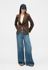Femme portant un blazer marron, un haut beige, un jean bleu à jambes larges, des tongs noires et une casquette bleu marine, debout les mains dans le dos.