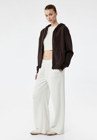 Brun zip-up hoodie, cropped hvid top og hvide vide bukser. Glatte teksturer; afslappet design; model poserer med hænderne i lommerne.