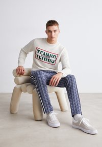 Pull crème en tricot avec logo "LACOSTE TENNIS" vert et rouge ; pantalon bleu à motifs avec bandes blanches ; baskets grises sur une chaise beige sculpturale.