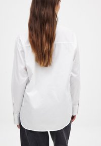 Weißes Langarmhemd mit geradem Schnitt, geknöpften Manschetten und abgerundetem Saum. Hergestellt aus glattem Stoff mit einem dezenten Glanz.