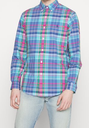 Hombre con una camisa de cuadros azul, rosa y verde de manga larga abotonada y jeans azul claro, de pie contra un fondo blanco.