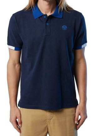 Mies, joka on pukeutunut tummansiniseen poolopaitaan, jossa on sininen kaulus ja hihansuut, sekä beiget housut, seisoo rentoin käsin.