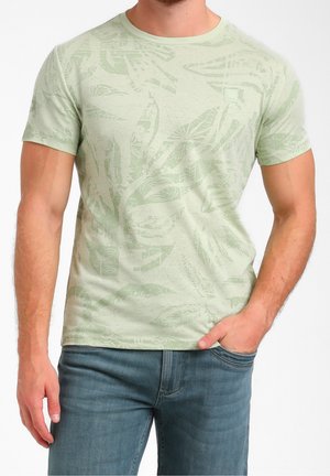 Man wearing light green short-sleeve T-shirt with subtle leaf pattern and blue jeans, one hand in pocket, on white background.