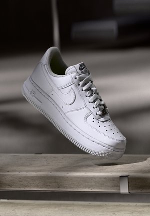 Baskets blanches Nike Air Force 1 avec lacets, semelle intermédiaire et bout perforé, affichées au-dessus d'une surface en béton avec un arrière-plan sombre flou.
