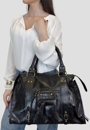 Donna con camicetta bianca e jeans che tiene una grande borsa in pelle nera con doppi manici e tasca frontale con cerniera.