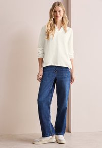 Hvid langærmet top med V-udskæring og samlede ærmer, parret med løstsiddende mørkeblå jeans og hvide sneakers. Neutral baggrund.