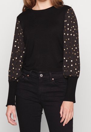 Frau trägt schwarzen Oberteil mit durchsichtigen langen Ärmeln mit goldenen Punkten, kombiniert mit hochtaillierten schwarzen Jeans.