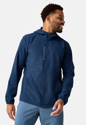 Moški se smeji in gleda v desno, nosi temno modro jakno s kapuco z logotipom "BROOKS" ter svetlo modre hlače na enobarvnem ozadju.