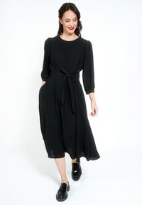 Žena u crnoj midi haljini s dugim rukavima i pojasom u struku, hoda prema naprijed noseći crne mokasine, smiješi se i gleda prema dolje.