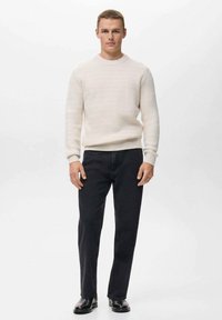 Suéter de punto color blanco roto con cuello redondo, combinado con pantalones vaqueros negros de pierna recta y zapatos negros, de pie contra un fondo liso.