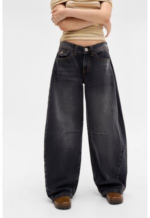 Personne portant un jean noir délavé à jambes larges, des chaussures marron et une chemise beige rayée à manches courtes, les bras croisés.