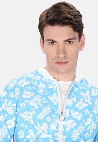 Jacheta bleu deschis cu fermoar, cu un model floral alb, având hibiscus și ananas, și o guler alb. Textură netedă și design casual.