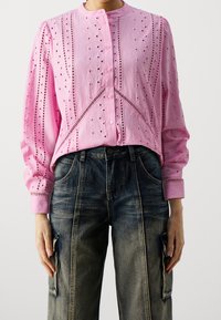 Camicia button-up rosa con dettagli in ricamo a occhio, maniche lunghe e colletto arrotondato, abbinata a pantaloni cargo in denim scuro con effetti strappati.