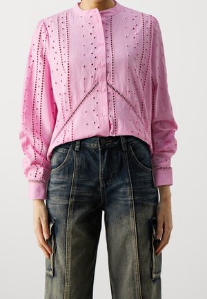 Camicia button-up rosa con dettagli in ricamo a occhio, maniche lunghe e colletto arrotondato, abbinata a pantaloni cargo in denim scuro con effetti strappati.