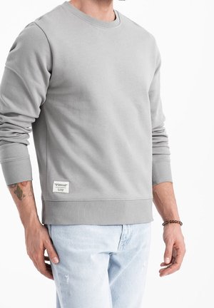 Mann trägt hellgraues Sweatshirt mit rundem Ausschnitt und hochgekrempelten Ärmeln, hellblaue Jeans und ein Perlenarmband am rechten Handgelenk.