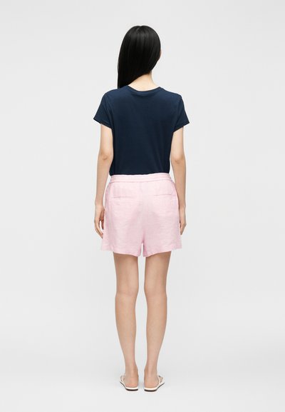 Γυναίκα με μακριά μαύρα μαλλιά που φοράει μπλε σκούρο μπλουζάκι, ανοιχτό ροζ σορτς με πίσω τσέπες και λευκά σανδάλια, στέκεται με την πλάτη προς τα εμπρός.