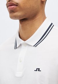 Κοντινό πλάνο ενός ατόμου που φοράει λευκό πόλο με μαύρες διπλές ρίγες στον γιακά και μικρό μαύρο κεντητό λογότυπο στο στήθος.