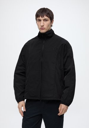 Homme debout de face, portant une veste noire zippée avec un petit logo sur la poitrine et un pantalon noir, sur un fond clair uni.