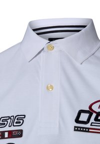 Weißes Poloshirt mit einem geknöpften Kragen, gelben Knöpfen und gesticktem Logo mit roten und schwarzen Akzenten sowie Flaggenmotiven auf der Brust.