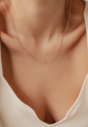 Silver necklace featuring a delicate chain with small round beads, resting on the collarbone, against a light textured fabric background.