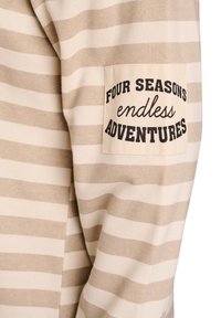 Camiseta de manga larga a rayas en beige y crema con un parche que dice "CUATRO ESTACIONES aventuras sin fin" en texto negro. Material de algodón suave.