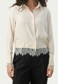 Cremefarvet bluse med krave, knaplukning foran og blondekant ved kanten og ærmerne. Glat stof med en delikat blomsterblonde detalje.