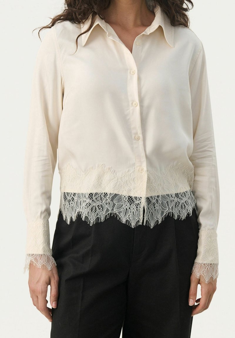 Cremefarvet bluse med krave, knaplukning foran og blondekant ved kanten og ærmerne. Glat stof med en delikat blomsterblonde detalje.