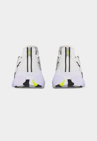 Ζεύγος λευκών αθλητικών παπουτσιών με μαύρες και νέον κίτρινες λεπτομέρειες, εμφανιζόμενα από πίσω σε απλό φωτεινό φόντο.