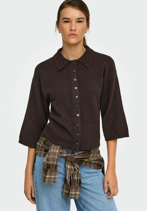 Vrouw draagt een donkerbruine trui met knopen en lichtblauwe jeans met een bruin geruit overhemd om haar middel gebonden, kijkt naar voren.