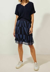 Granatowa bluzka z dekoltem w kształcie V i krótkimi rękawami, w zestawieniu z granatową spódnicą z geometrycznym wzorem, z falbaniastym wykończeniem. Nosi z białymi trampkami.