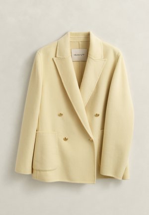 Cream double-breasted wool blazer with notch lapels, two front patch pockets, and gold buttons, hanging against a light background.