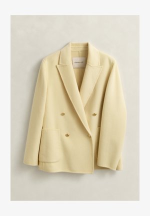 Blazer en laine crème à double boutonnage avec revers à encoche, deux poches plaquées sur le devant et boutons dorés, suspendu sur un fond clair.