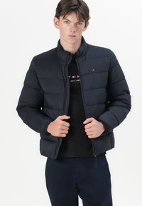 Marineblå pufferjakke med horisontalt quiltet design, høy krage, full glidelås og minimal merking på venstre bryst. Glatt tekstur.