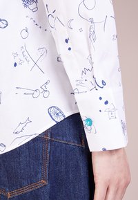 Camicia bianca con motivi astratti blu, dotata di un bottone turchese sul polsino. Jeans di denim visibili nella parte inferiore.