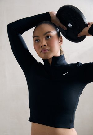 Svart atletisk crop-top med dragkedja i kragen, försedd med en Nike-logga, bärs av en person som lyfter en viktdisk över huvudet.