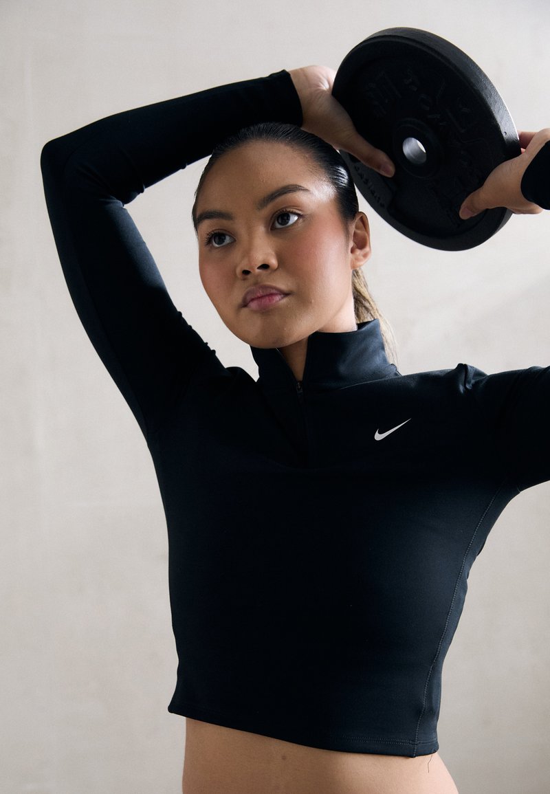 Musta urheilu crop top, jossa on vetoketjukaulus ja Nike-logo, henkilön päällä, joka nostaa painolevyä päänsä yläpuolelle.