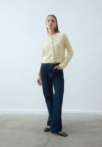 Jasnożółty kardigan o fakturze z okrągłym dekoltem i złotymi guzikami, zestawiony z ciemnoniebieskimi prostymi dżinsami i czarnymi balerinami.