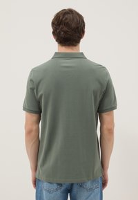 Hombre con camiseta tipo polo de manga corta de color verde oliva liso y vaqueros azules, de pie de espaldas sobre un fondo liso y claro.
