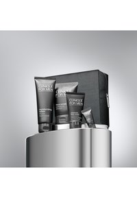 Ett set med fyra svarta hudvårdstuber märkta "Clinique for Men" i en zippered tygpås, placerade på en cylindrisk silveryta.