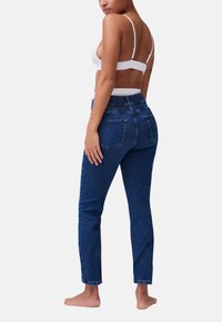 Jeans in denim scuro a vita alta, con una silhouette aderente, tasche posteriori visibili e un orlo pulito, abbinati a un top reggiseno bianco.