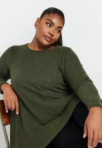 Grön stickad långärmad tröja med rund halsringning och avslappnad passform. Texturerad tyg, sidovalvsdetaljer och mjuk drapering.