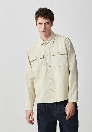 Junger Mann trägt eine beigefarbene Jacke mit Knöpfen und Überschlagstaschen sowie dunkle Hosen, steht vor einem schlichten hellgrauen Hintergrund.