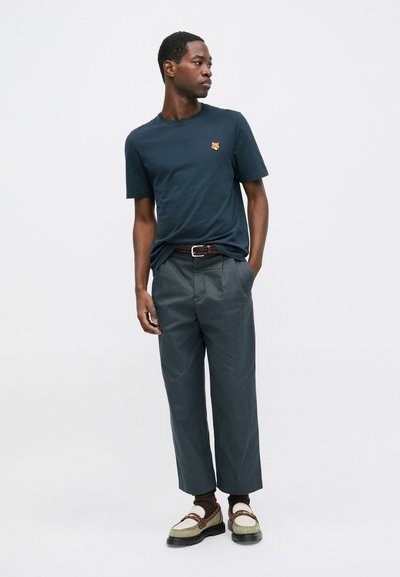 T-shirt bleumarin cu un mic logo de vulpe portocalie, asociat cu pantaloni largi gri și mocasini verzi cu accente maro.