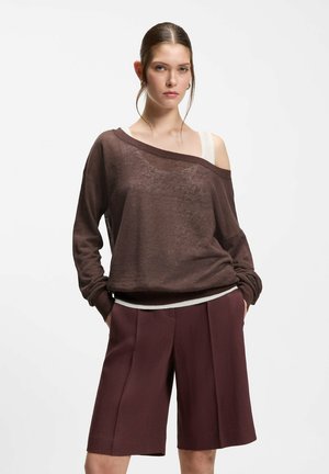 Young woman with brown loose off-shoulder sweater over white top, burgundy knee-length wide-leg shorts, standing with hands in pockets.