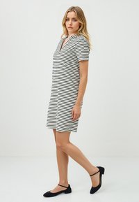 Una donna sta in profilo, indossando un vestito corto a righe e scarpe basse nere, con i lunghi capelli che scendono sulla spalla su uno sfondo semplice.