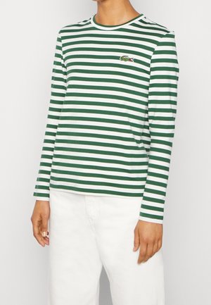 Persoon die een groen-wit horizontaal gestreept shirt met lange mouwen draagt met een klein krokodillenlogo en een witte broek.